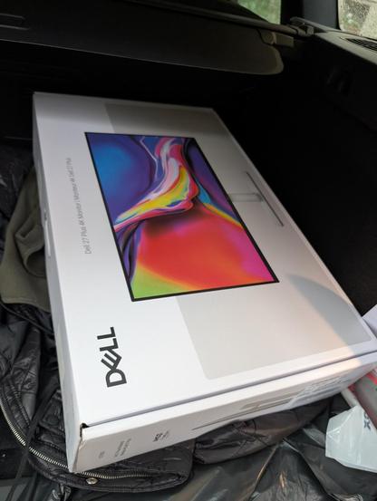 A Dell monitor box, specifically the Dell 27 4K Monitor (model S2725QS), is placed in the back of a vehicle. The box features a colorful image of the monitor screen and is resting on a black jacket or blanket. The vehicle's interior and part of the window are visible in the background.