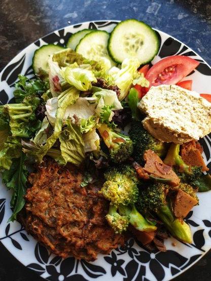 A plate with the meal of a baked fritter made from root vegetables; Stir fried homemade seitan, broccoli & pod peas; Homemade hummus; Side leaf salad w/ tomato & cucumber.