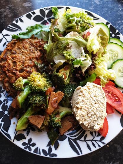 A plate with the meal of a baked fritter made from root vegetables; Stir fried homemade seitan, broccoli & pod peas; Homemade hummus; Side leaf salad w/ tomato & cucumber.