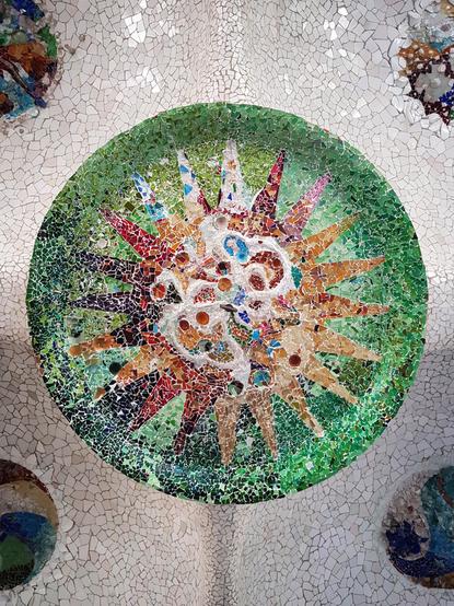 A close-up, upward view of a circular, sun-shaped mosaic on the ceiling of Park Güell in Barcelona, Spain. The mosaic is primarily green with a multi-coloured sunburst design in the centre, made from broken ceramic tiles. The surrounding ceiling is white mosaic.
