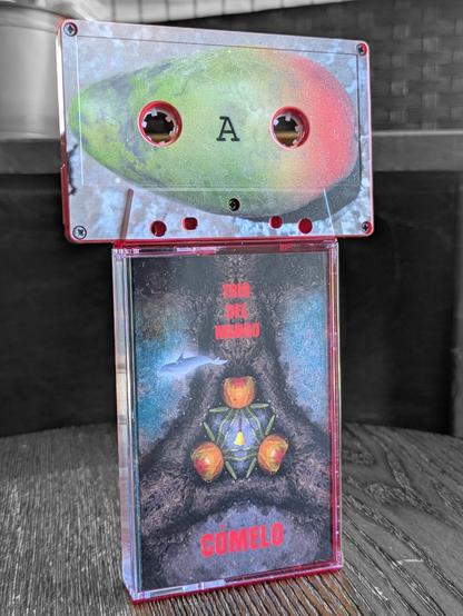 the physical cassette tape and case + cover art of Trio del Mango "Cómelo" displayed with cassette sitting on top of the case. the cover art is a depiction of a tree with 3 Mangos at its base, a bell and a dolphin. the tape itself  looks like a Mango.