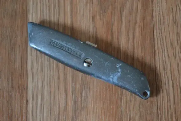 A worn gray metal knife with the blade retracted lying on a wooden table. Stamped on the side is the branding: Stanley 99 E.