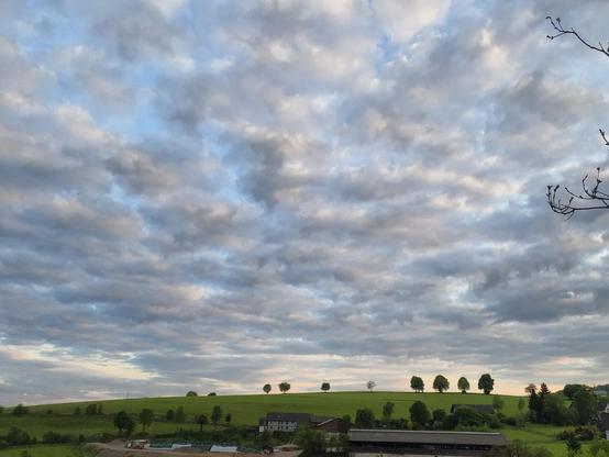Grauweiße Wolken liegen wie getupft über dem grünen Hang, angeleuchtet von der Abendsonne. Am Horizont bilden einige Bäume eine Linie. Davor ist das Dach eines Bauernhofes zu erkennen.