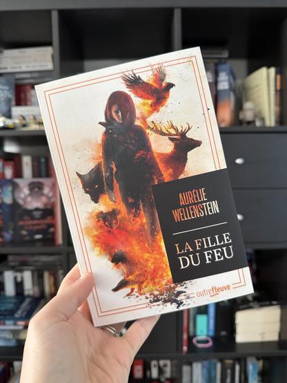 Livre pris en photo devant une bibliothèque. 
La couverture de La fille du feu montre une jeune fille marchant dans les flammes entourée d’animaux sauvages
