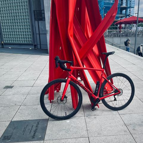 Ein rotes Rennrad lehnt an einer ebenso roten Skulptur aus metallenen Streben, die aus dem Boden in Richtung Himmel führen.