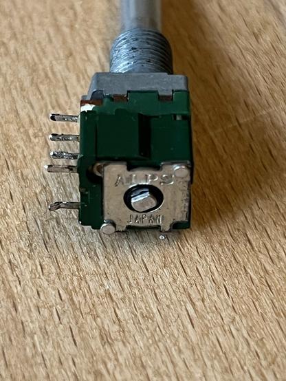 Drehpotentiometer mit grünem Gehäuse und metallener Front und Achse. Zu sehen sind die Anschlüsse und die Beschriftung auf der Rückseite ALPS Japan. Detailaufnahme der Rückseite mit einer Metallplatte.
