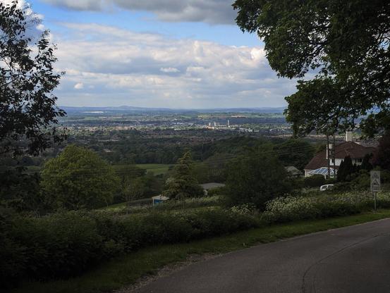 A picture of a city taken off a road on a higher level of terrain. Signs for a sleep slope and house can be seen close to the camera, with the cityscape of Gloucester further out with hills in the background. Trees are obscuring part of the image.