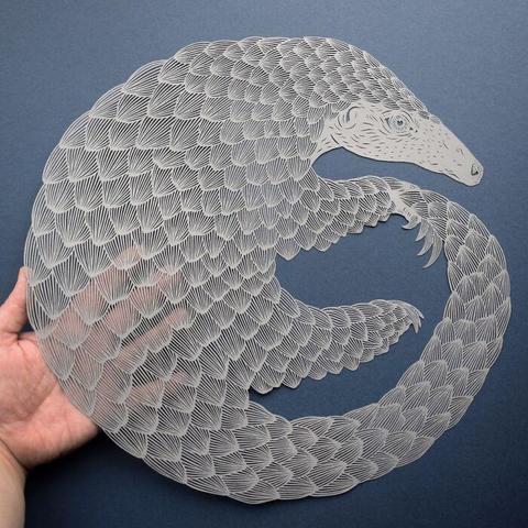 The image features a detailed paper cutout of a pangolin, a scaly mammal, arranged in a circular form. The pangolin is depicted with intricate lines representing its scales, creating a textured appearance. The cutout is held by a hand, providing a sense of scale. The background is a solid dark blue, which contrasts with the light-colored paper, making the pangolin stand out prominently. The pangolin's head is positioned at the top of the circle, with its body curving around to form the circular shape. The tail is visible, extending from the bottom right, and the overall design is symmetrical. The craftsmanship is evident in the precision of the lines and the delicate nature of the cutout.

Provided by @altbot, generated privately and locally using Ovis2-8B