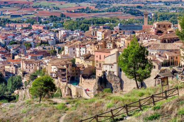 Aquesta imatge mostra una vista panoràmica de Conca, Espanya. Les cases tradicionals de color terra s'aferren a vessants rocosos, creant un paisatge urbà únic i impressionant. L'estil arquitectònic és clarament històric, amb cobertes de teula i balcons sobresortint. La imatge captura una barreja d'arquitectura antiga i moderna, amb edificis més moderns visibles al fons, en contrast amb el caràcter històric de les cases antigues de la ciutat. Les formacions rocoses naturals que recorren la ciutat afegeixen un element dramàtic al paisatge. El verd dels arbres i el marró sec del sòl circumdant proporcionen una atractiva gamma de colors.