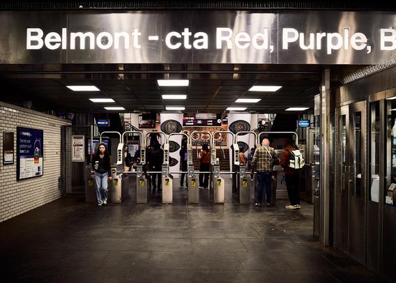 The image shows the entrance of the Belmont CTA station in Chicago. Stainless steel turnstiles stand in a row with people passing through them. Overhead, a sign reads 'Belmont - cta Red, Purple, Brown Line'. The lighting is bright with tiled walls and floors