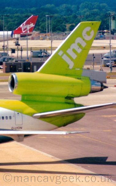Close-up of the tail and centre engine of a 3 engined jet airliner taxiing from right to left.
The plane's fuselage is white, with a bright, lime green tail with diagonal, white "JMC" titles on the top section.
The registration "G-LYON" is on the lower rear fuselage in black.
Tall lighting stands are dotted around the background, with the red and white tail of another jet aircraft at the top left.