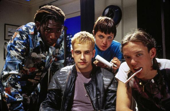 The image shows four young individuals grouped closely together in an indoor setting. The person on the left has dreadlocks and wears a colorful, patterned shirt while leaning slightly forward. The person in the center has short blond hair, a black leather jacket, and a calm expression. To their right is an individual with short dark hair, wearing a blue top and holding a white object in their hand. The person on the far right, wearing a white and black patterned shirt, has their hair tied back and a striped stick, possibly candy, protruding from their mouth. The background includes a window showing a nighttime cityscape, a partial clock on the wall, and framed pictures.