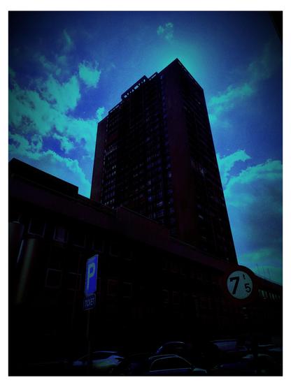 Das Bild zeigt eine hoch aufragende Wohngebäude in einer städtischen Umgebung. Die Perspektive ist von unten nach oben gerichtet, was die Höhe des Gebäudes betont. Der Himmel ist blau mit vereinzelten weißen Wolken, die einen kontrastreichen Hintergrund bilden. Das Gebäude selbst ist dunkel und hat viele Fenster, die in regelmäßigen Abständen angeordnet sind. Im Vordergrund sind ein Parkplatzschild mit der Aufschrift "P" und "TICKET" sowie ein Verkehrszeichen mit der Beschriftung "7t 5" zu sehen. Parkplätze sind sichtbar, und die gesamte Szene ist in einem düsteren, blaugrünen Farbton gehalten, was eine düstere Stimmung vermittelt.