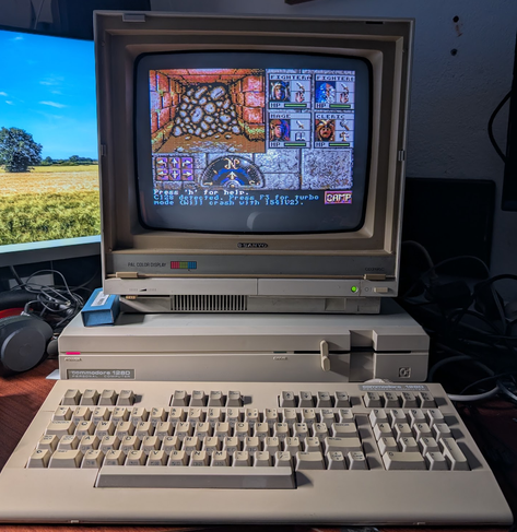 A Commodore 128D: On the main unit that looks like an Amiga 1000 with a 5,25" floppy drive, there's a Sanyo "PAL Color Display", in front of the computer is it's seperate keyboard. On the monitor there's the opening scene of the Commdore 128 enhanced modern port of Eye of the Beholder.