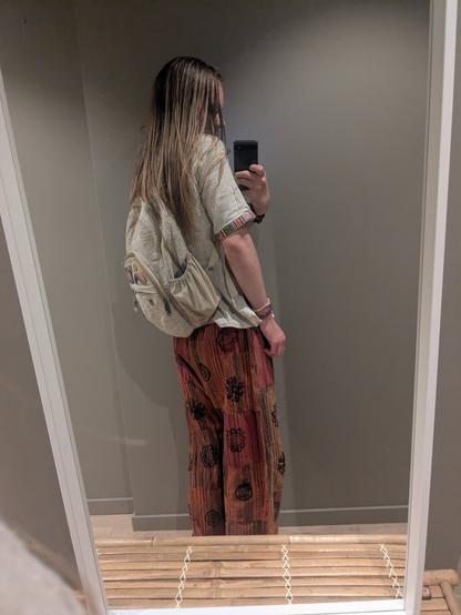The image features a single individual standing indoors in a personal space. The person is facing a mirror, holding a phone to capture a reflection. They are wearing a casual outfit with a backpack.