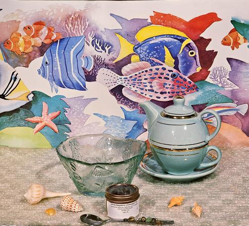 Tea tableau celebrating sea life. Against a watercolor backdrop of ocean fishes are a green glass bowl embossed with clamshells, sea stars, and sea horses, along with real seashells, a spoon embellished with sea glass and pebbles, an aqua green tea for one service, and Champagne Raspberry white loose-leaf tea from Indulge Littitz.