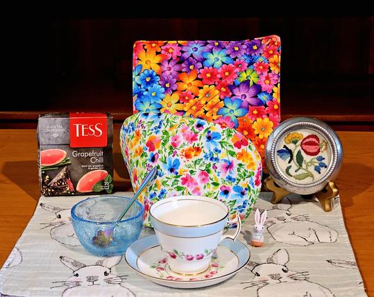 Springtime tea tableau in bright flowery colors with a floral and rabbit theme, vintage china, vintage glass, antique embroidery, and a vintage miniature wooden bunny ornament. The tea is Grapefruit Chill black tea from Tess Teas, flavored with grapefruit and mint.