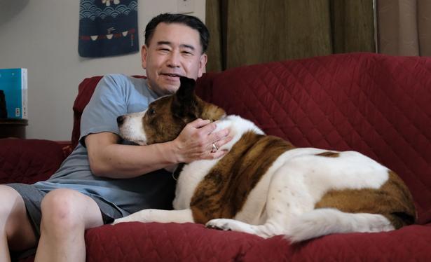 A rescued man in a blue T-shirt sitting on a couch with a wine-colored cover, holding a white and brown rescue dog lying sideways with his head just under the man's face