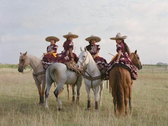 four women sit on horseback in a prairie, wearing colorful dresses and wide-brimmed hats