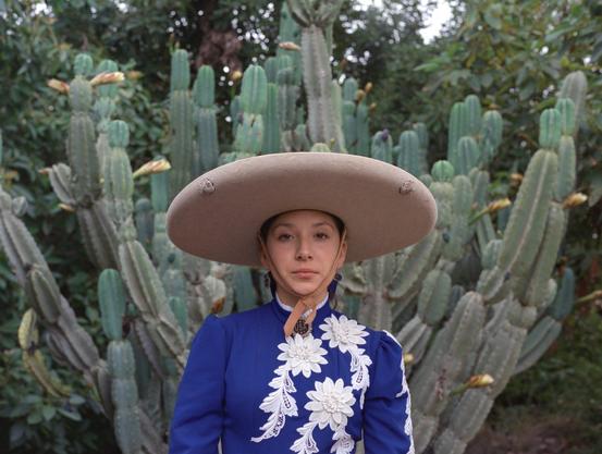 a woman stands in front of a cactus, wearing a blue-and-white top and a wide-brimmed hat