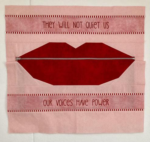 A small quilt top featuring a giant pair of pieced red lips held closed by an actual silver zipper, shown in full on a design wall. Above and below are several borders that stretch from side to side, made from two alternating peach fabrics set off with red and white striped accents. The area between the two accents is machine embroidered in a fun red font - above reads “They will not quiet us” and below reads “Our voices have power”.