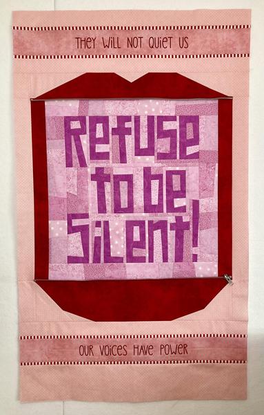 The same zipped lips quilt top as in the first photo, only the zipper has been unzipped and the quilt is much longer. Though the top and bottom borders are still visible, a wide center section now shows as well, so that the quilt top resembles an open mouth. Inside the ‘mouth’ are wonkily pieced letters reading “Refuse to be Silent!” The letters are sewn from a pinkish purple fabric and set on a scrappy purplish pink background.