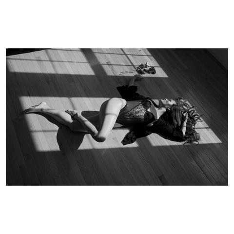 female model laying on a wooden floor under sun light beaming from a window directly on her and her left hand reaching for a bunch of flowers