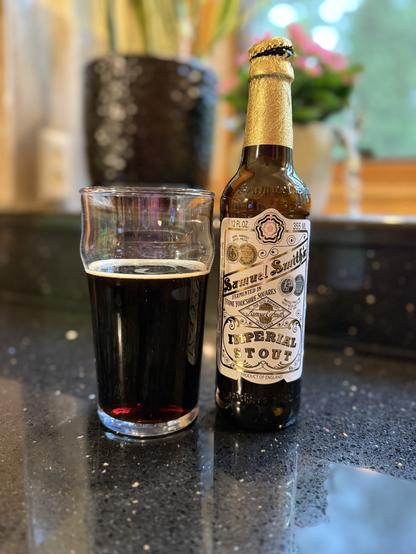 A pint glass of dark beer next to a brown bottle with a bright gold neck foil and a white and gold label