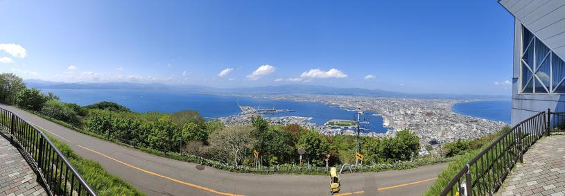 A panoramic photograph taken from the top of a modestly tall mountain. You see the long narrow strip of land that composes Hakodate leading out from the island in the shape of a fan, flanked by ocean on both sides