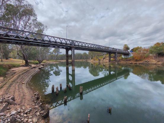 The iron-frame, wood-deck John Foord bridge is reflected in the gum tree lined Murray River, under a low overcast sky.