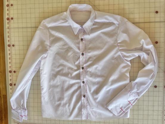 A white button front shirt with French cuffs lays flat on a gridded cutting table. The shirt is obviously roughly sewn and is covered in magenta marker notations.