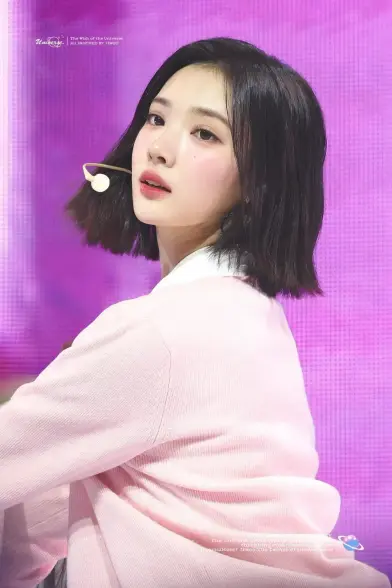 The image features Kpop idol Jiwoo with shoulder-length dark hair, wearing a light pink sweater over a white collared shirt. The background is a vibrant purple with a subtle grid pattern. The person is wearing a small white microphone headset, suggesting they might be performing or speaking. The text in the image reads "The Wish of the Universe" and "All Inspired by Jiwoo," indicating a theme or inspiration related to the person. The overall color scheme is soft and pastel, with the pink sweater and purple background creating a harmonious and visually appealing contrast.

Provided by @altbot, edited by @jemmesedi