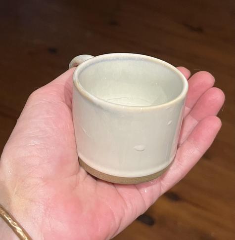 Smol off white stonewear mug with an unglazed strip of brown at the bottom sitting in the palm of my hand. Hardwood floor below. Mug capacity is 3oz.