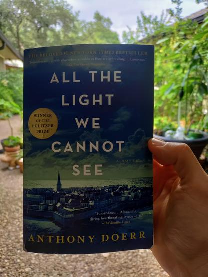 Paper back book ALL THE LIGHT WE CANNOT SEE being held by a Caucasian person's hand while apparently outdoors.  The book cover displayed the Pulitzer prize winner medal.