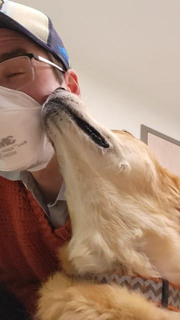 Selfie of a young whiteish man and an orange dog. The dog has turned her head upwards to lick his face. The man is wearing an n95 ask and the dog's tongue is on it, his eyes are closed out of suprise. It is very cute. He is wearing a blue hat and a pumpkin orange sweater his mom made him.