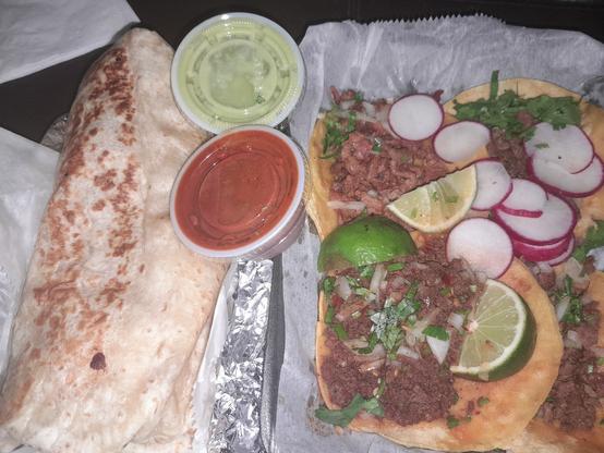 A large burrito, red and green salsa containers, and a to-go container of Mexican street tacos with lime wedges.