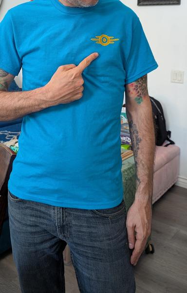 Photo of a dude wearing a blue shirt, pointing at a small logo embroidered on the chest in gold and yellow