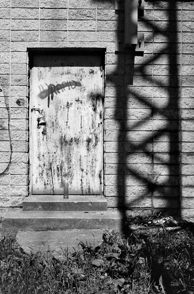 The black and white image shows a weathered metal door with peeling paint and graffiti, set in a brick wall. A single candle stands on the concrete step in front of the door, and the shadow of a metal structure casts a diagonal pattern across the wall and ground.