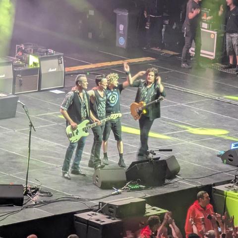 Los cuatro músicos de Stone Temple Pilots están sobre el escenario después de la actuación. Dos de ellos sostienen guitarras, una de color claro y otra de color marrón oscuro. El que toca la guitarra marrón está de pie con los brazos alrededor de los hombros de los otros dos. El músico de la guitarra clara está de pie entre los otros dos. Todos ellos parecen estar sonriendo o felices. Al fondo, se ve el público y equipo de sonido.