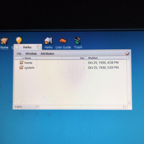 The image shows a computer screen displaying a file explorer window with a blue background. The window is titled "Haiku" and contains two folders named "home" and "system." The "home" folder was last modified on October 25, 1930, at 4:58 PM, while the "system" folder was modified at 5:09 PM on the same date. The file explorer window has tabs labeled "File," "Window," and "Attributes," with columns for "Name," "Size," and "Modified." The desktop icons visible at the top of the screen include "home," "Haiku," "User Guide," and "Trash." The overall appearance suggests an older operating system interface, possibly Haiku, with a vintage aesthetic.