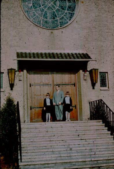 A man and two girls who might be twins standing before a set of large wooden doors at the top of a stone staircase. Above the door is a tiled or possible corrugated overhang. Above the overhang is a rose window that appears to have a sword pattern. On either side of the doorway are two large amber glass and iron lanterns. Further out are two small stained glass windows that are slightly smaller than the lamps.