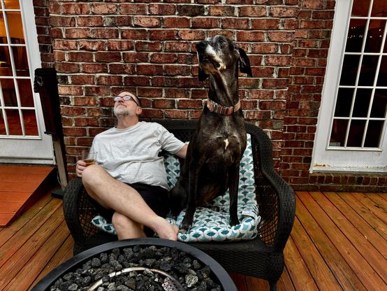 An old, slow, grey bearded man sitting on a patio love seat beside his old, slow, grey muzzled Great Dane friend.