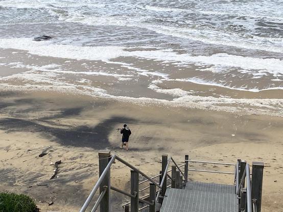 A barefooted person wearing a black long sleeved top and shorts using a metal detector on the beach after the storms. He's got another piece of equipment resting on his left shoulder (maybe a shovel?). He's not far from the long flight of stairs that lead down to the beach. This is in the aftermath of intense storms and heavy rain brought by an East Coast Low. The light sand of the beach has lots of dark stains. The waves have pushed dirty cream foam onto the beach and the water looks a tad grungy.