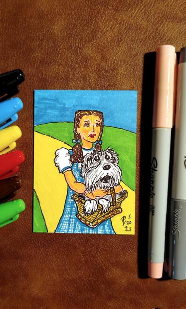Marker drawing of Judy Garland as Dorothy from the film "The Wizard of Oz." Instead of Toto, the basket she's carrying contains Sprocket the dog from "Fraggle Rock." The drawing is signed with my "RV" signature glyph and dated 5-20-25. The black, turquoise, yellow, red, brown, green, pink, and grey markers used to draw the image rests on the table next to the card.