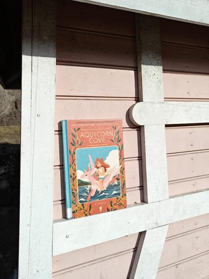 Kuvassa Aquicorn cove -kirja kuvattuna vaaleanpunaista seinää vasten.