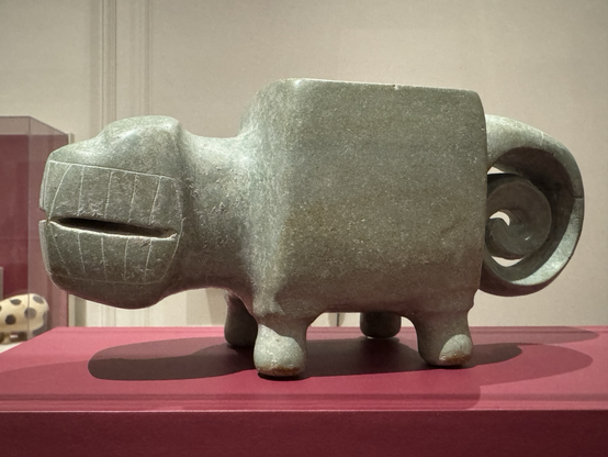 photo of mortar #1 (larger one) on display at museum in side profile: greenstone sculpture in the form of a quadruped with square body, head with large mouth full of teeth, short legs, and a spiral tail