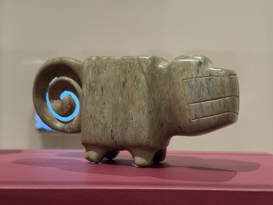 photo of mortar #2 (smaller one) on display at museum in side profile: greenstone sculpture in the form of a quadruped with square body, head with large mouth full of teeth, short legs, and a spiral tail