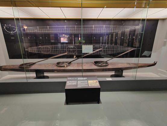 A photo of a museum exhibit. It is a two person kayak made from wood and hide. It sits behind glass with a large info plaque Infront of it.
