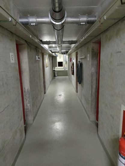 Blick in den kargen Beton-Hauptgang des Bunkers. Rote Vorhangtüren zweigen ab. Notausstieg am Ende.