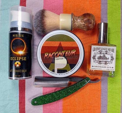 (Le Chat Mistral description)

The image showcases a collection of grooming items arranged on a striped background. There is a black bottle of aftershave balm labelled "Eclipse" on the left. Next to it, a round tin of shaving soap named "Raconteur" sits with a shaving brush on top. A bottle of cologne labelled "Chatillon Lux" is positioned to the right. Below these items, a green-handled straight razor is placed. The items are neatly organised, suggesting a traditional shaving setup.